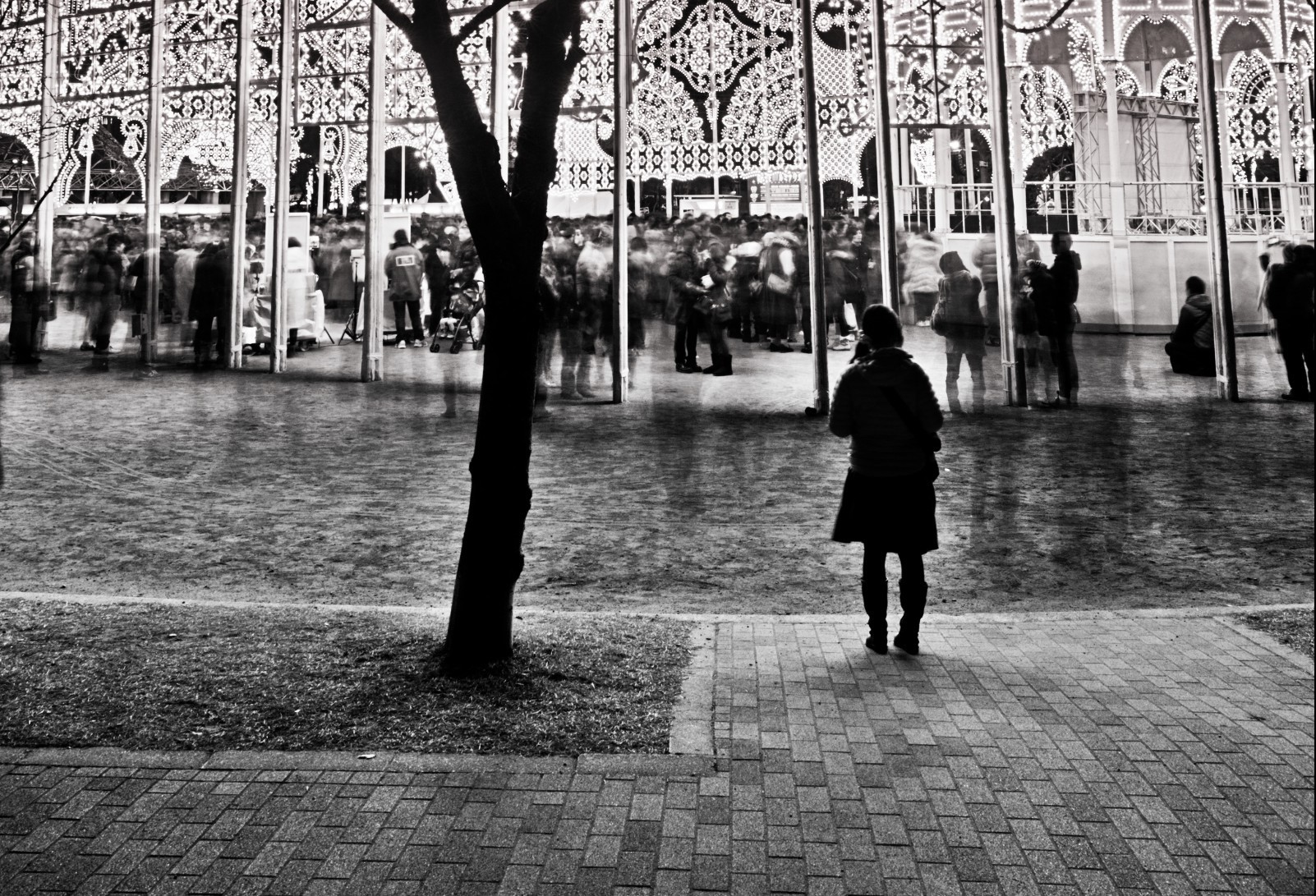 Image of a black and white night scene captures a festive illumination event. a solitary figure stands on a brick pathway, observing a crowd gathered under intricately lit arches. the contrast between the dark silhouette and bright lights highlights the vibrant, bustling atmosphere of the urban celebration