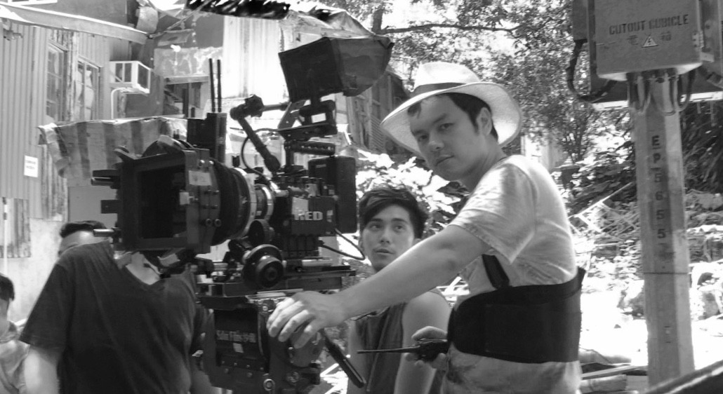 Image of a film crew on-set managing a professional red camera in an outdoor shooting location. the scene captures the effort of filmmakers in action, with one crew member wearing a wide-brimmed hat to shield from the sun. the background suggests an urban setting with
