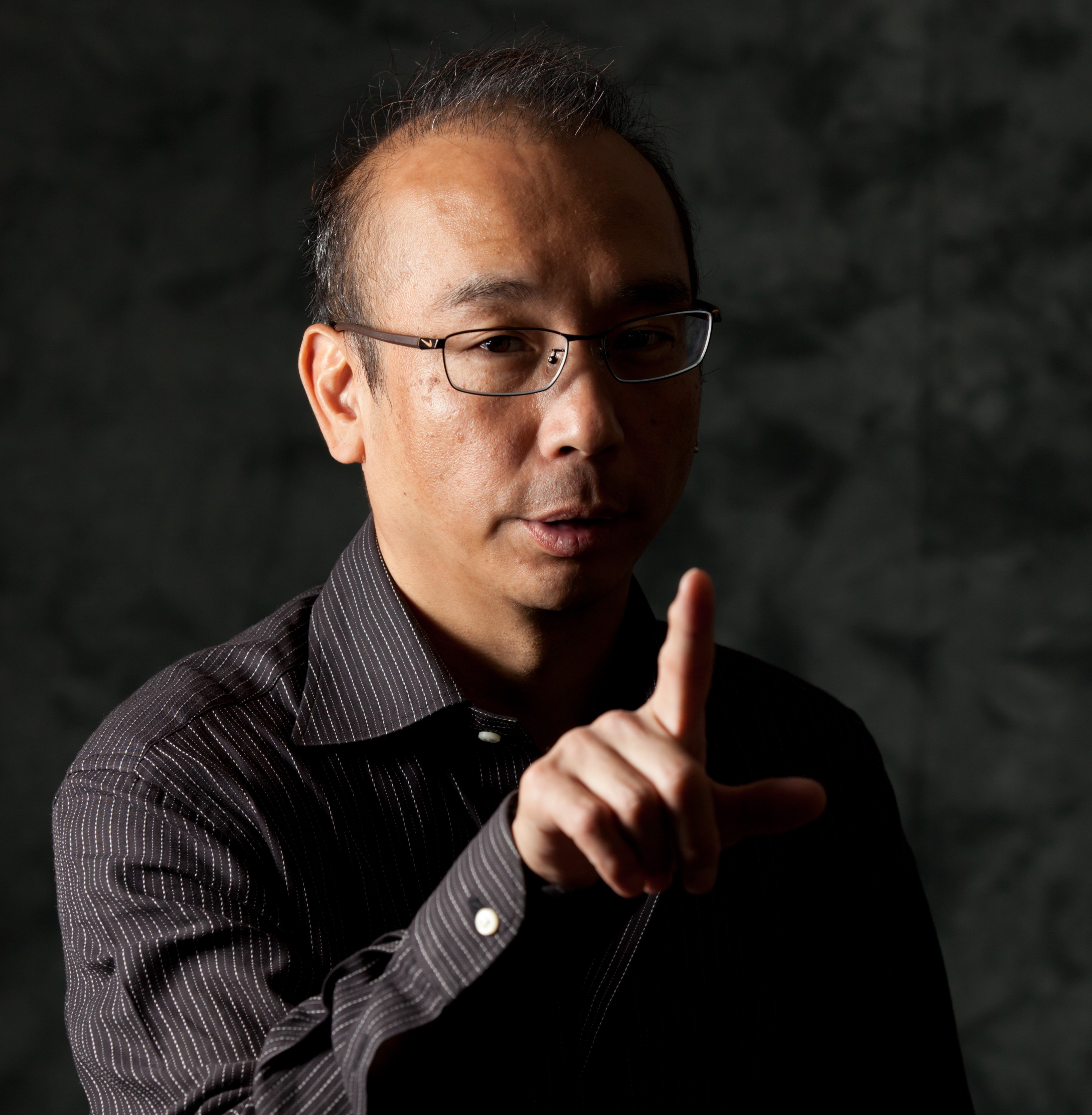 Image of a person with glasses wearing a striped shirt makes a gesture with their finger against a dark background. the dramatic lighting emphasizes a serious expression, creating a powerful portrait. keywords: portrait photography, dramatic lighting, gesture, glasses, striped shirt, dark background.