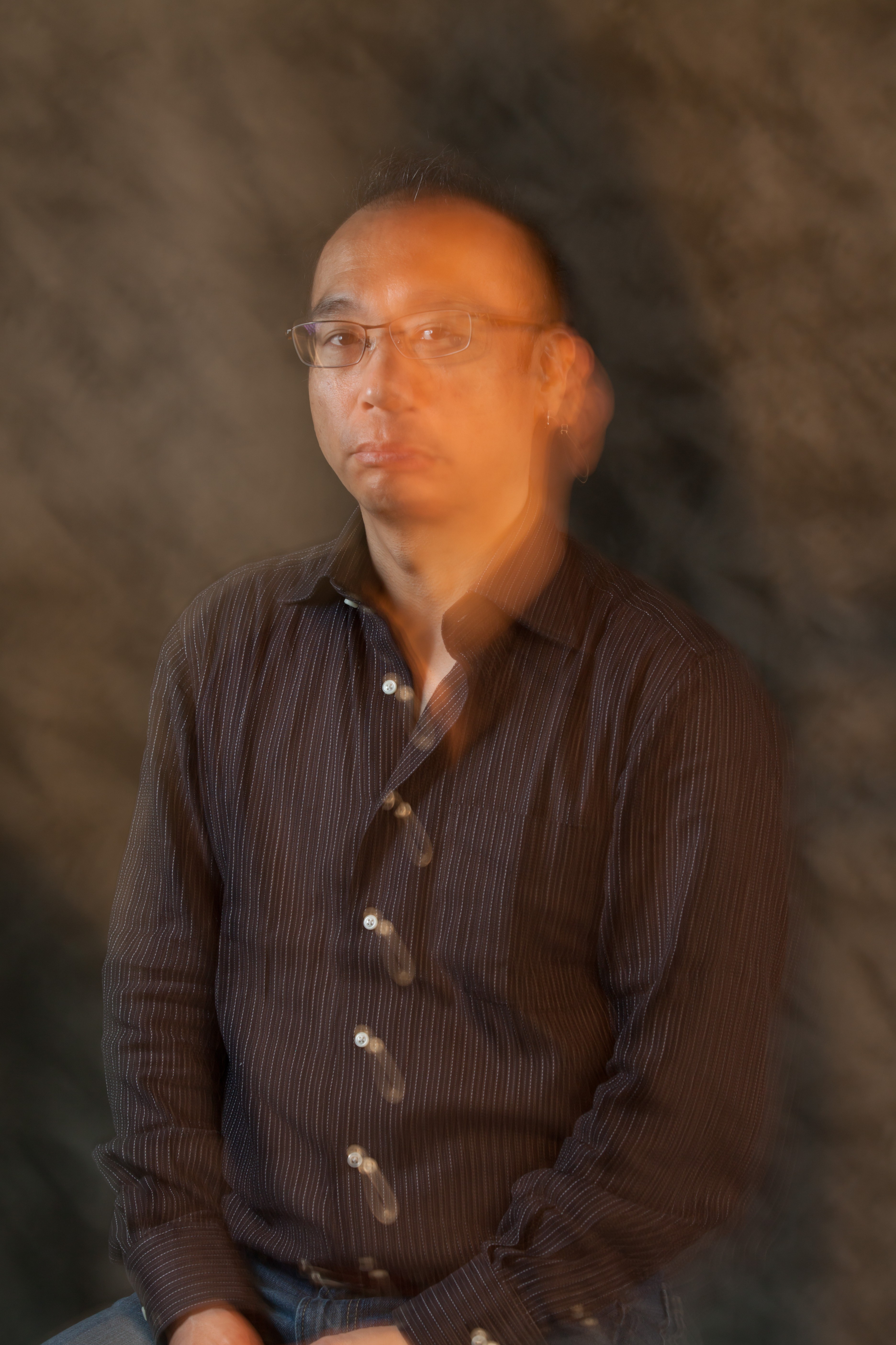 Image of a blurred portrait of a man in a striped shirt and glasses, set against a textured dark background. his head appears in motion, creating a dynamic visual effect. this intentional motion blur adds an artistic, ethereal quality to the image. ideal keywords: