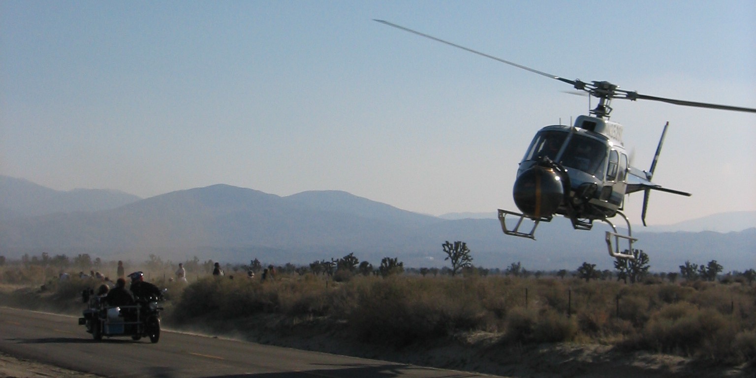 Image of helicopter flying low over a desert road, capturing a dynamic scene with a motorcycle and sidecar on the pavement below. the rugged desert landscape and mountains in the background enhance the adventurous atmosphere. ideal for keywords like "desert road adventure," "