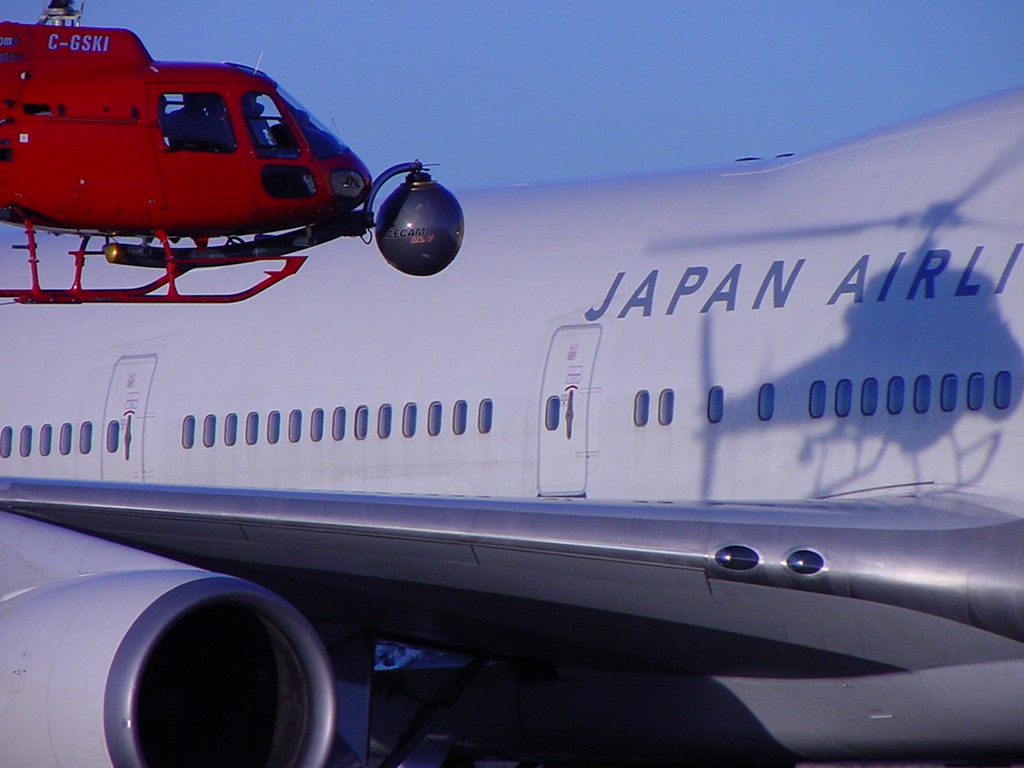 Image of a red helicopter hovers close to a japan airlines boeing 747 airplane. the photo captures the aircraft's side with "japan airlines" prominently displayed, highlighting aviation operations or airshows. the helicopter, equipped with filming or observation gear, suggests a professional