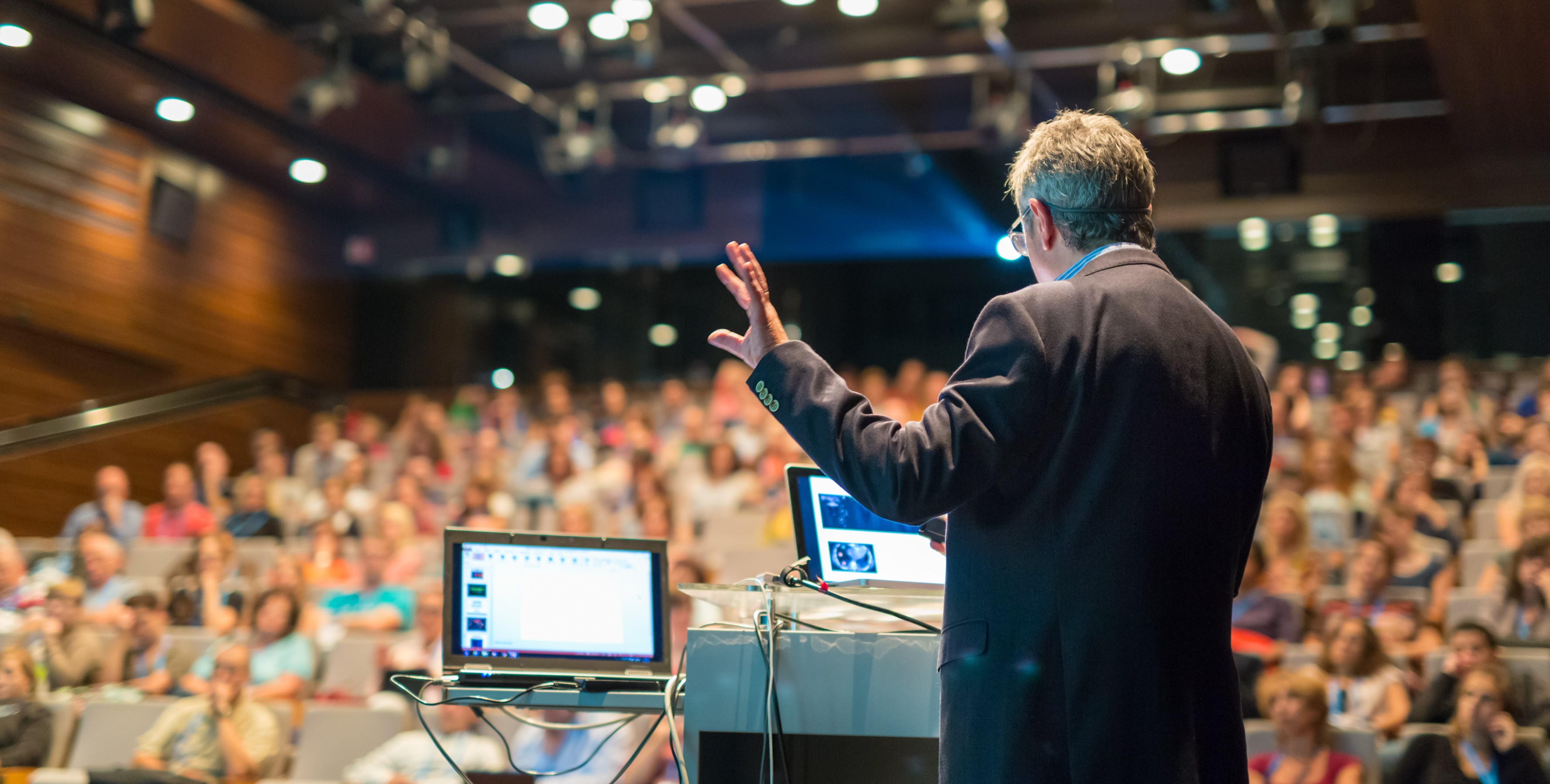 Image of a speaker delivers a presentation to a packed audience in a modern conference hall. the scene captures the dynamic interaction between the presenter and the engaged attendees, with a focus on the speaker's hand gestures and the use of technology with laptops. this setting is ideal