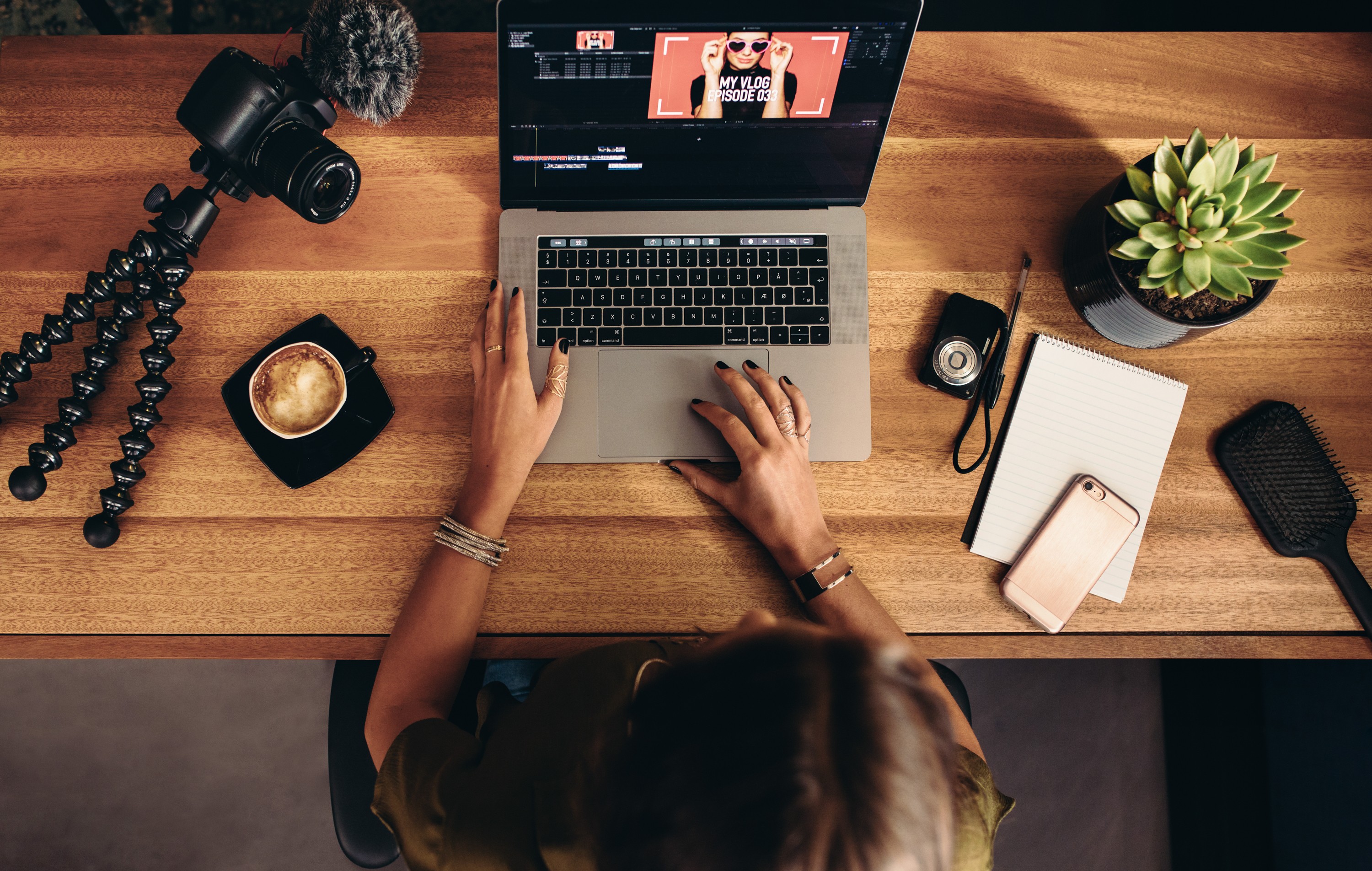 Image of a vlogger's workspace featuring a person editing a video on a laptop with "my vlog episode 03" on the screen. the desk is equipped with a camera on a flexible tripod, a cup of coffee, notepad, smartphone, and