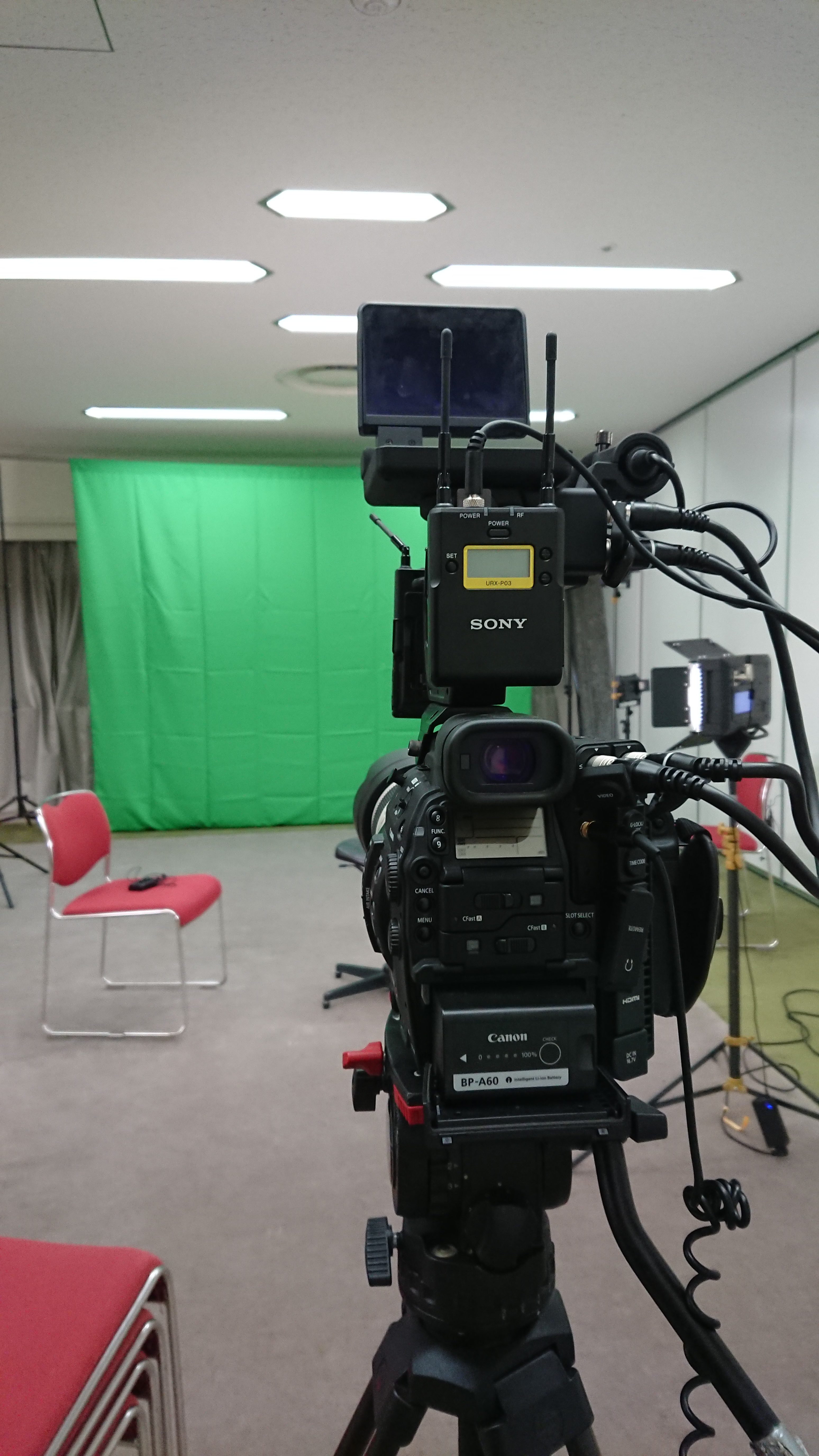 Image of a professional video production setup in a studio featuring a digital camera on a tripod aimed at a green screen backdrop. bright studio lights illuminate the scene, while red chairs are positioned for interviews or presentations. this setup is ideal for video editing, chroma key