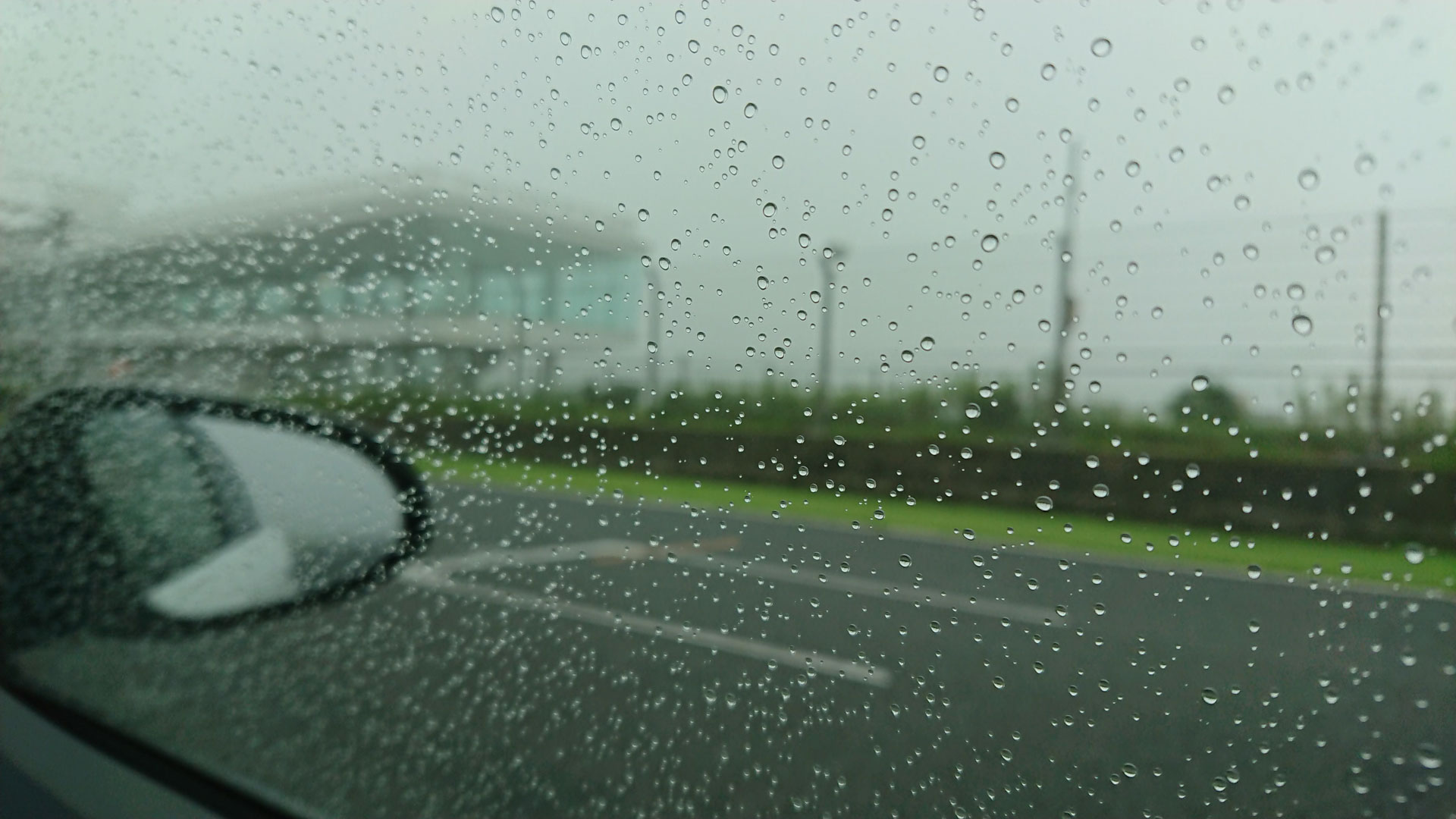 Image of rainy day captured from a car window, featuring water droplets on the glass and a blurred view of a roadside landscape. the overcast sky and wet road create a moody atmosphere, perfect for a stormy weather theme. ideal for keywords like rainy