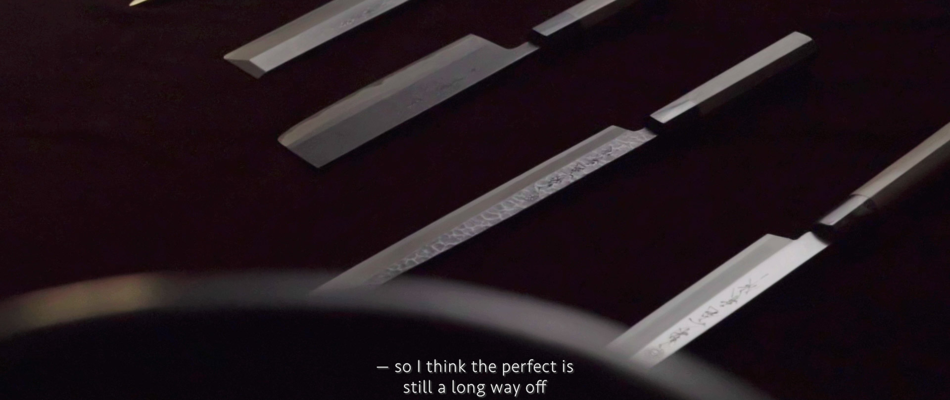 Image of this image shows a close-up of finely crafted japanese knives laid out in a row on a dark surface. the blades, displaying intricate patterns and details, highlight traditional japanese craftsmanship. subtitles at the bottom provide a contemplative statement about the pursuit of perfection