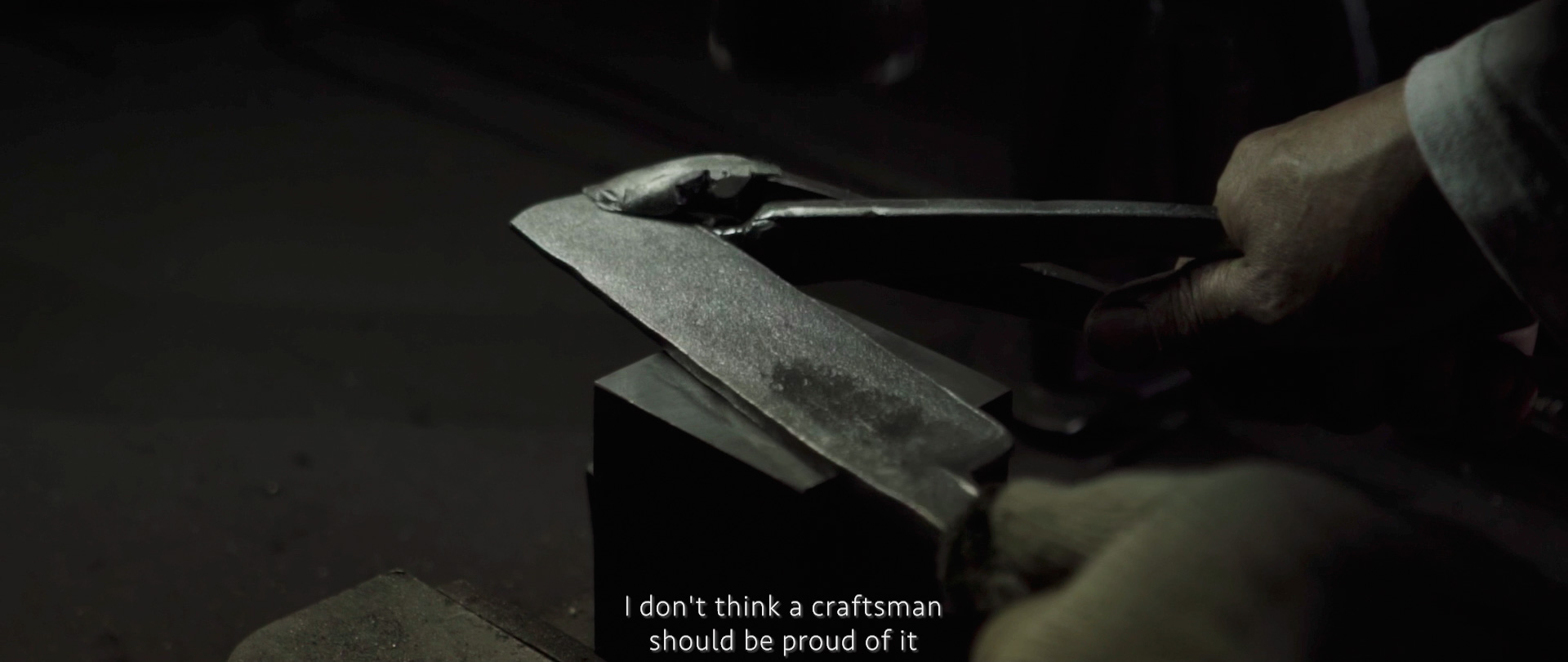 Image of the image depicts a metalwork scene focused on a craftsman shaping a tool or weapon on an anvil. the close-up view highlights the texture of the metal and the craftsman's steady hand. the setting is dimly lit, emphasizing the traditional black
