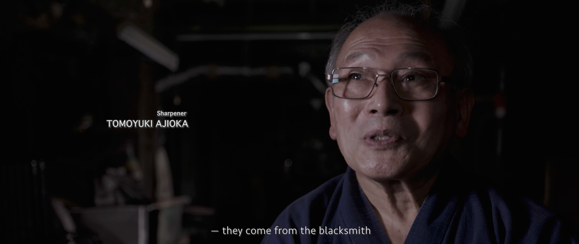 Image of the image captures a portrait of a knife sharpener discussing his craft, with the subtitle "they come from the blacksmith." the scene is dimly lit, emphasizing the authenticity and traditional ambiance of the setting. this captures the essence of japanese craftsmanship and