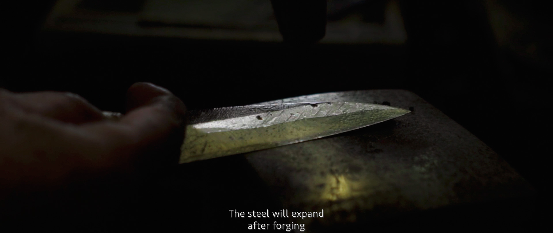 Image of a close-up of a hand holding a freshly forged steel blade in a dimly lit blacksmith workshop. the blade rests on an anvil, showcasing raw craftsmanship. text overlays the image, stating "the steel will expand after forging," highlighting the traditional