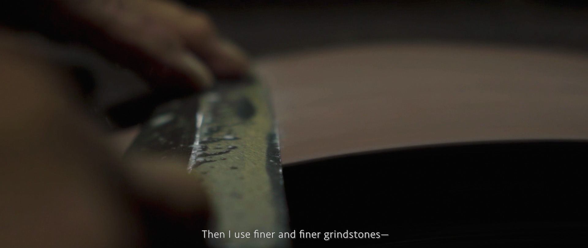 Image of close-up image of a craftsman sharpening a blade on a grindstone, showcasing precision and skill in metalwork. the focus on the hands and the blade highlights traditional knife sharpening techniques. the text overlay reads, "then i use finer and finer grind