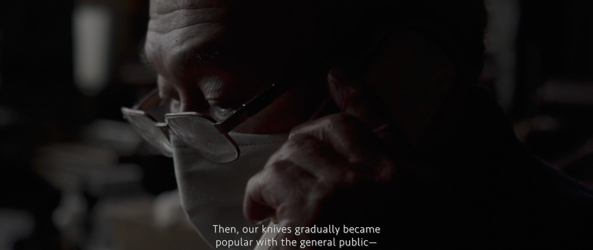 Image of the image depicts a close-up of an elderly person wearing glasses and a face mask, engaged in a thoughtful moment. the lighting is low, creating a contemplative and intimate atmosphere. subtitles reveal a conversation about knives gaining popularity. this scene could relate