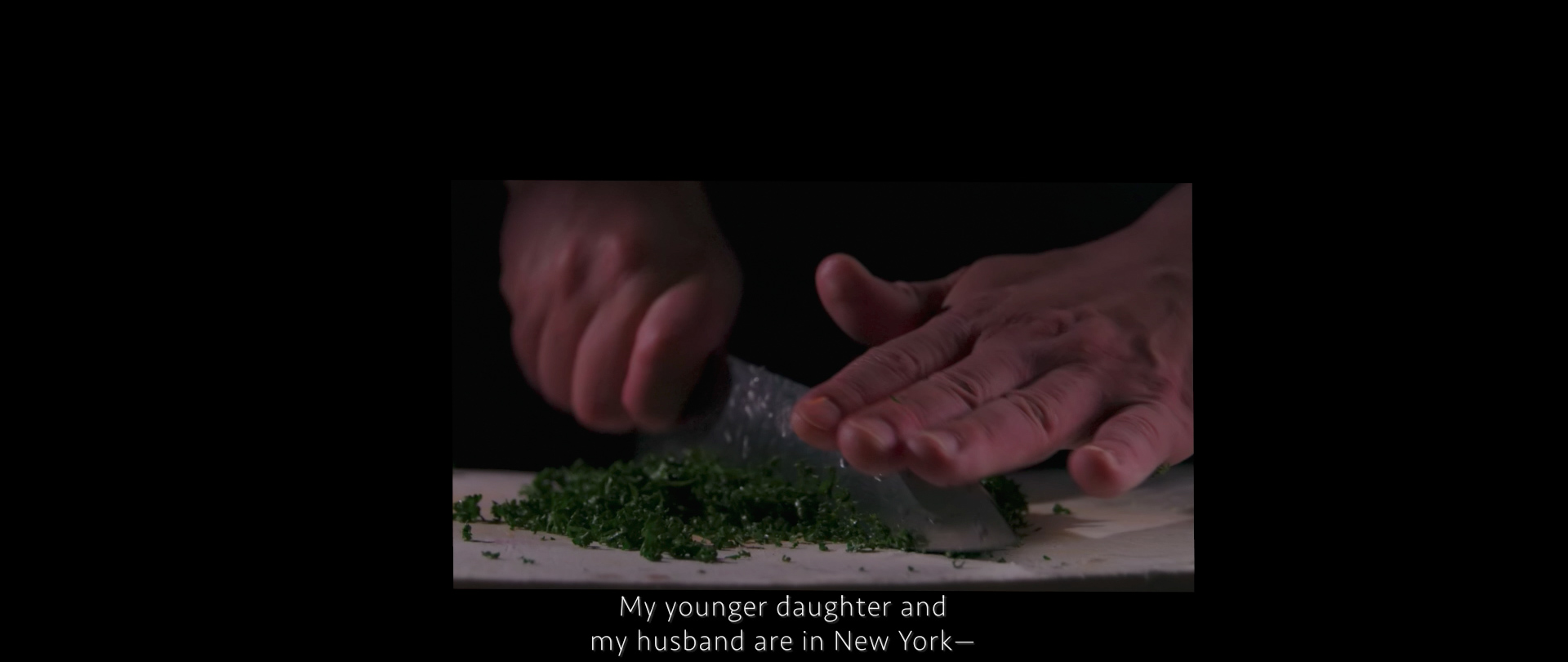 Image of a close-up shot of hands skillfully chopping fresh herbs on a cutting board, set against a dim background. the scene suggests an intimate kitchen atmosphere, focused on culinary preparation. subtitles indicate a conversation about family in new york, adding a personal touch