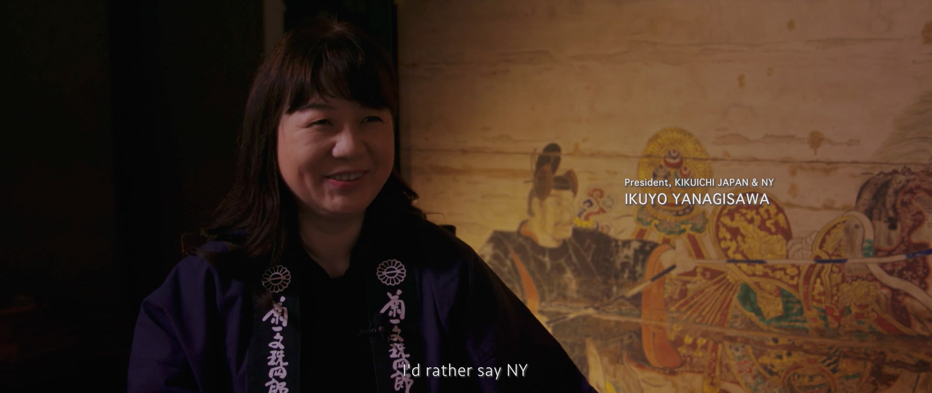 Image of in this image, a person is smiling while sitting against a backdrop featuring traditional japanese art. the setting looks cultural and intimate, enhancing the theme of japanese heritage and art. the person is identified as the president of kikuichi japan & ny. the