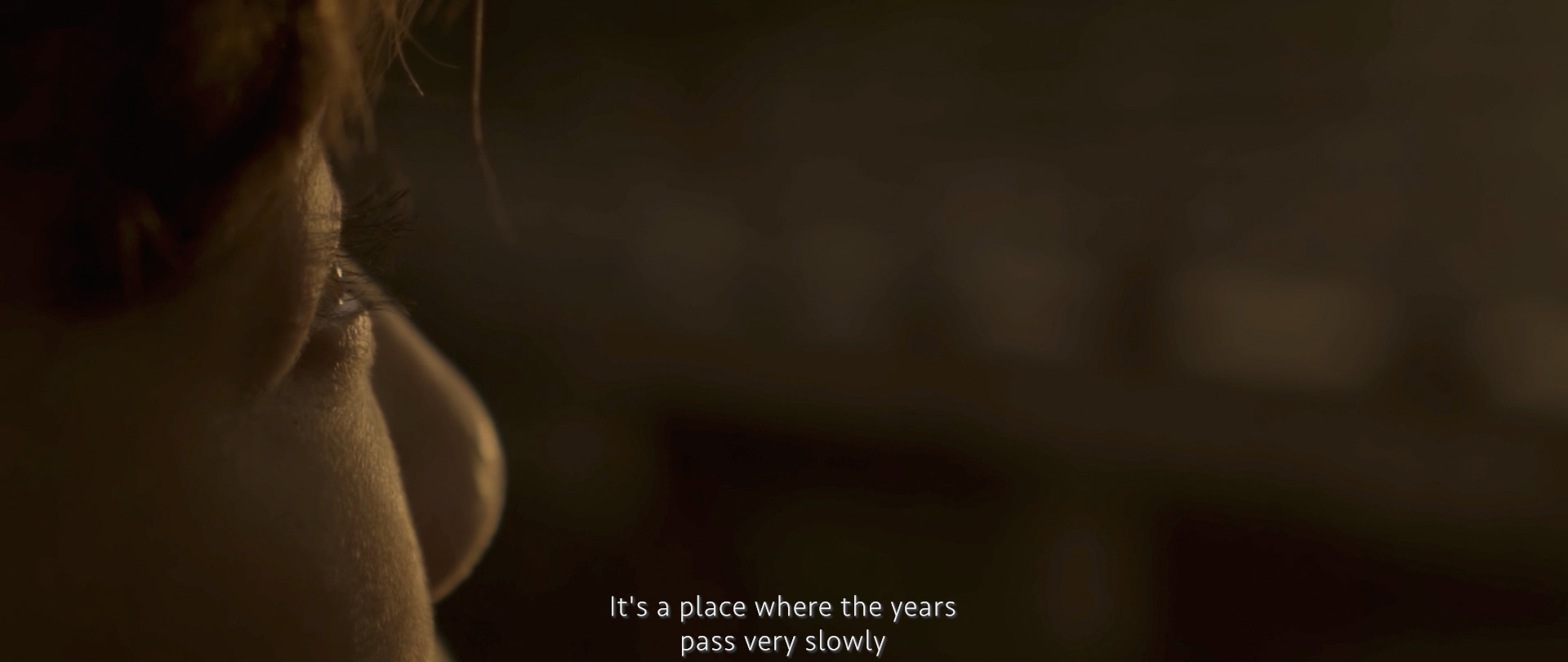 Image of the image features a close-up of an individual's face focusing on the eye, captured in warm, ambient light. the background is softly blurred, adding depth and introspection. the text overlay reads, "it's a place where the years pass very slowly,"