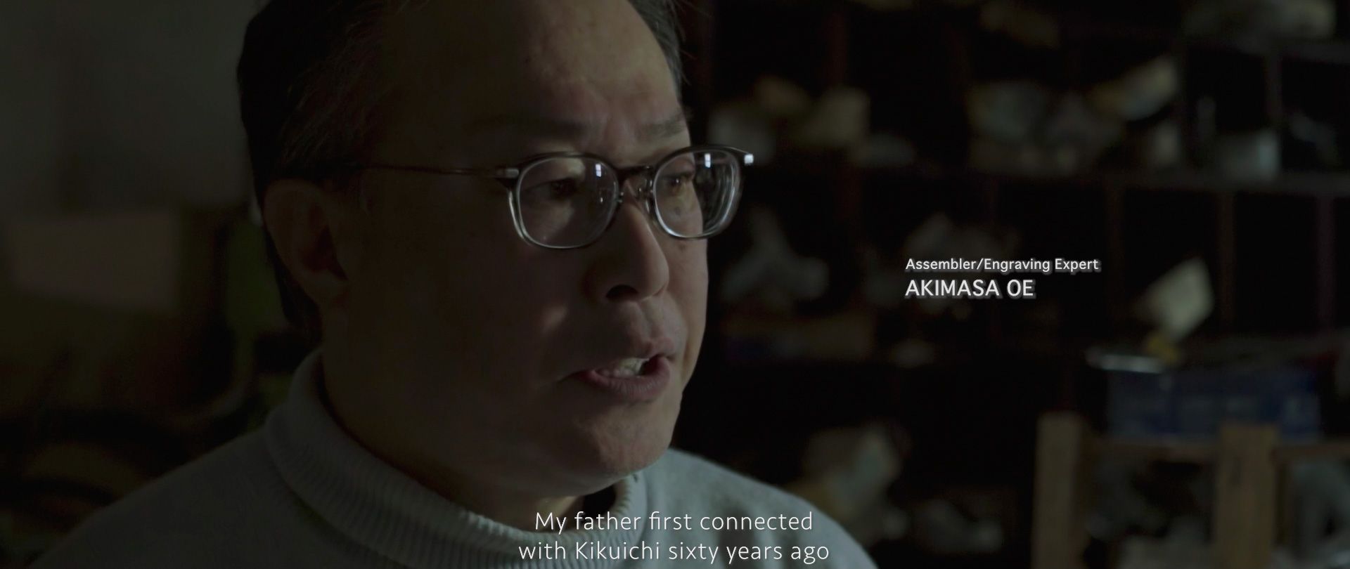 Image of akimasa oe, an assembler and engraving expert, is depicted in a dimly lit setting, conveying a personal story about his father's connection with kikuichi from sixty years ago. the image focuses on oe's expression, emphasizing his expertise