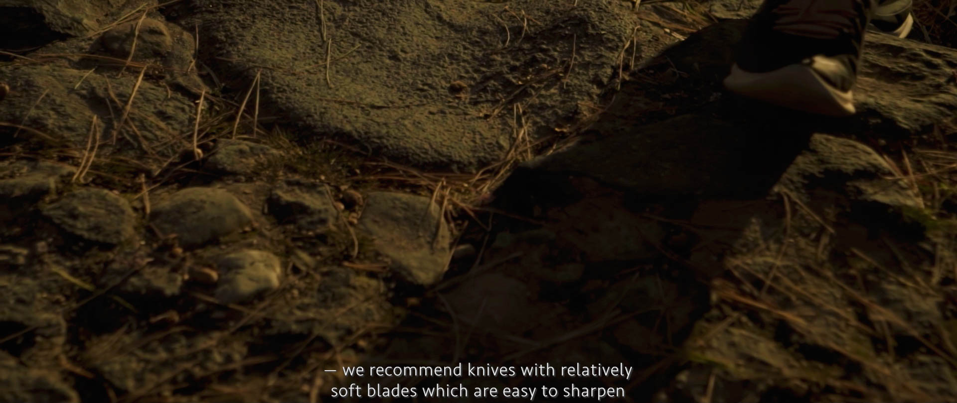 Image of a person walks on a rocky, forest-like terrain covered with dry pine needles. the scene is dimly lit, suggesting early morning or late afternoon. the text overlay recommends knives with soft blades for easy sharpening. keywords: rocky terrain, pine needles,