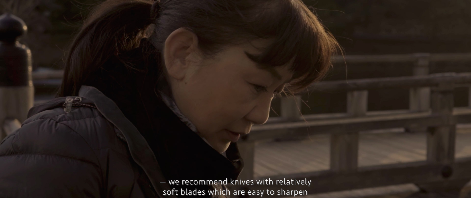 Image of a person in a winter jacket is outdoors, framed by a wooden railing, during a serene sunset. the scene suggests a peaceful moment, possibly near a park or waterfront. subtitles on the image discuss the recommendation of using knives with relatively soft blades for
