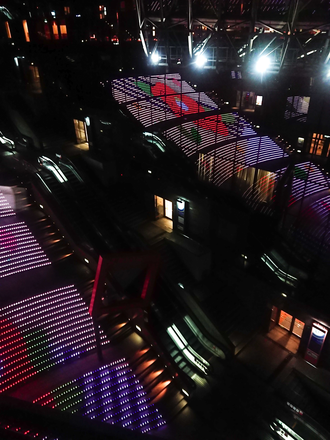 Image of nighttime view of a cityscape featuring a striking architectural display in a japanese urban setting. the scene includes dynamic led light installations on stairs, illuminated in vibrant colors like red, purple, and white. the modern design highlights the use of technology and creativity