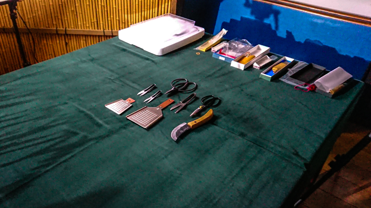 Image of the image shows a neatly organized collection of bonsai tools on a green tablecloth, ideal for bonsai enthusiasts. scissors, pruning shears, and other specialized implements are visible, set against a rustic bamboo backdrop, creating a serene and traditional gardening