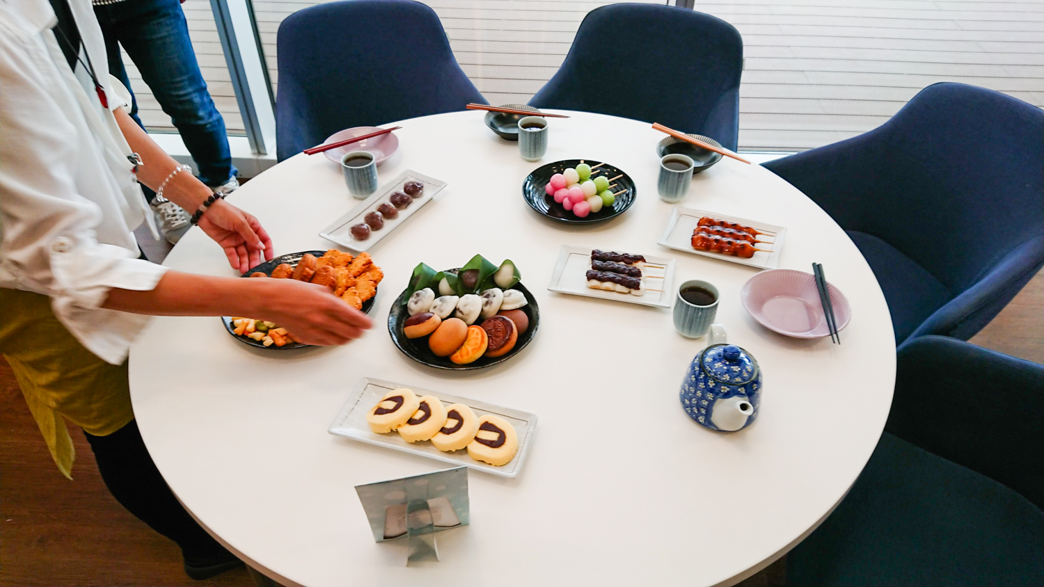 Image of a round table is elegantly set with a variety of traditional japanese sweets, including dango, manju, and other confections. the table is surrounded by dark blue chairs, and a person is arranging dishes. cups of green tea and a blue te