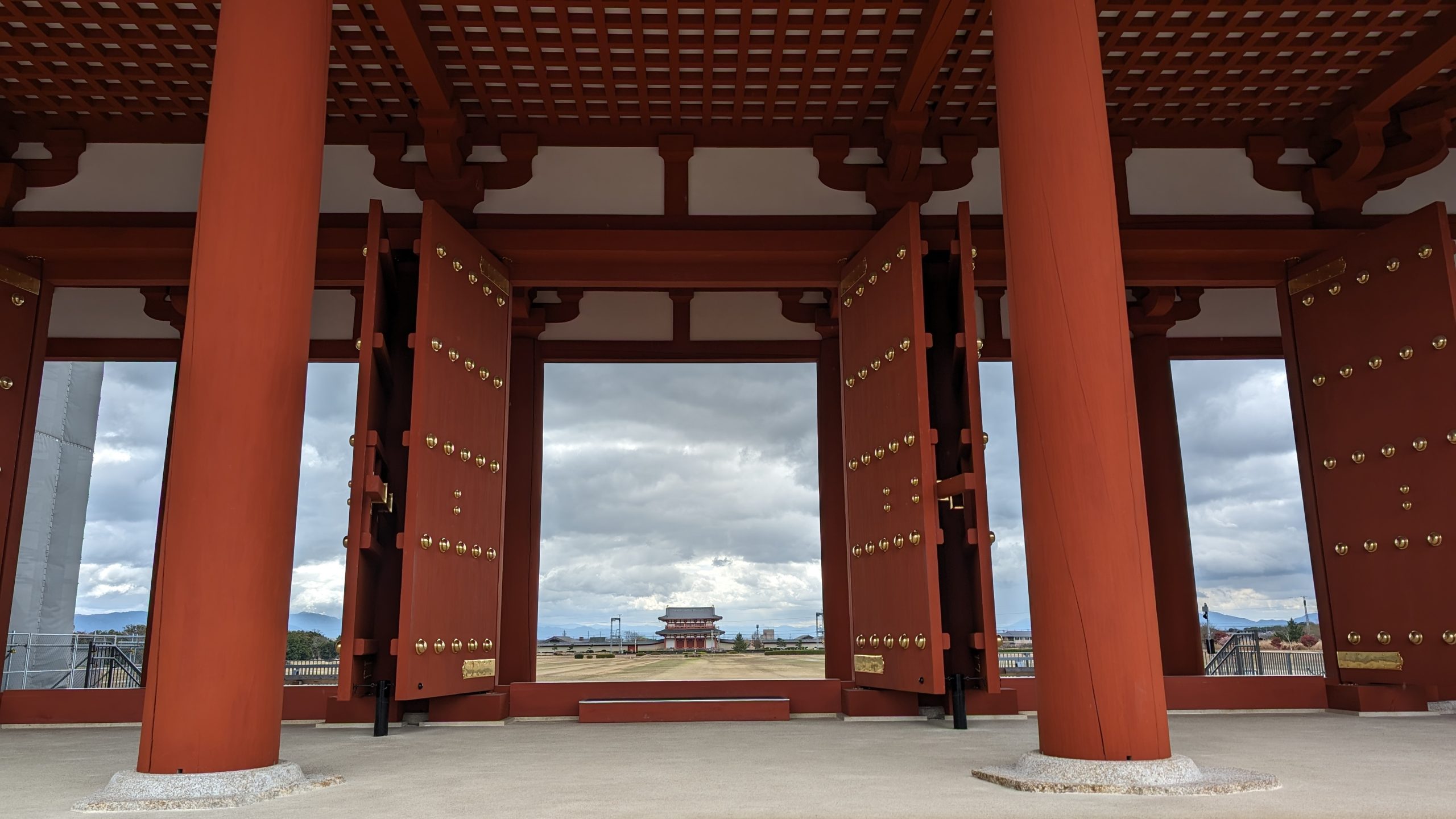 Image of majestic view through the grand wooden gates of heijō palace in nara, japan. this historical site features traditional japanese architecture with large red pillars and gold accents, opening to a scenic view of the ancient palace complex under cloudy skies. perfect for