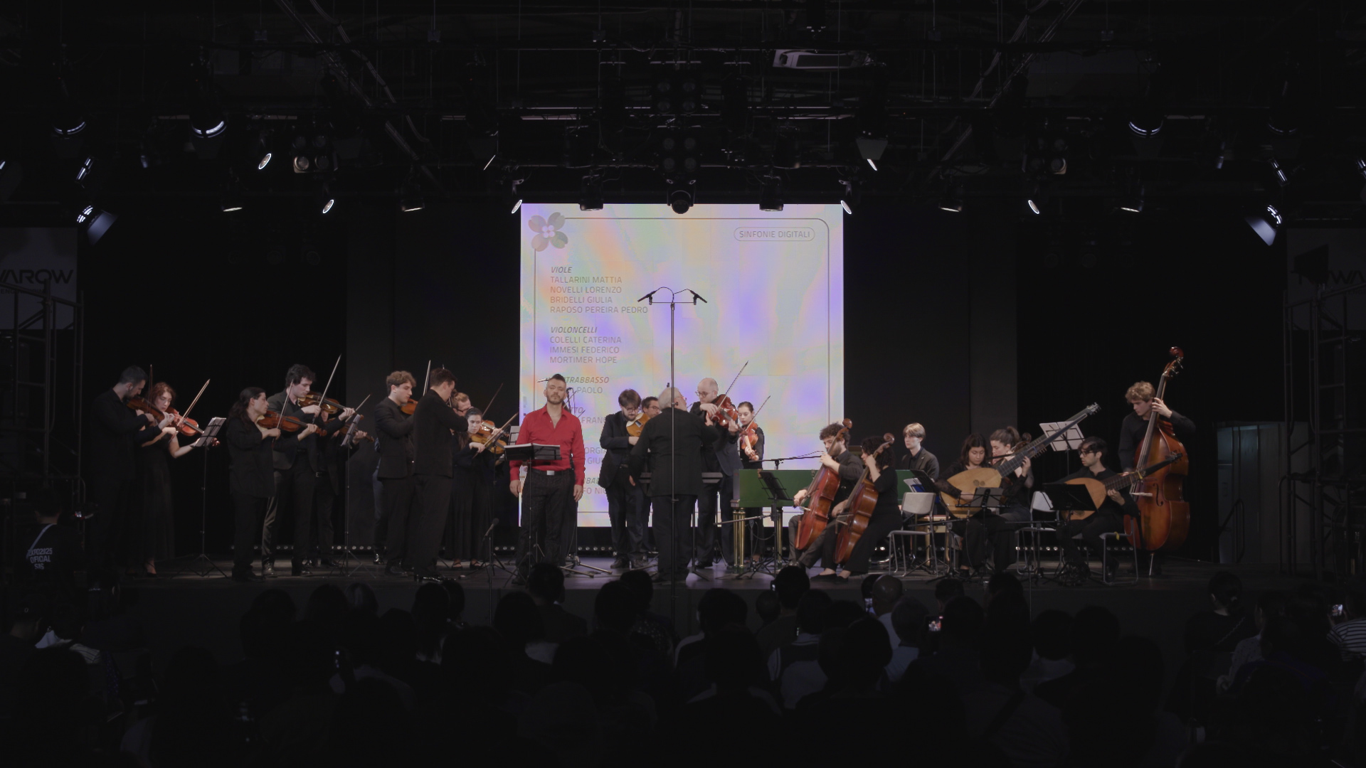 Image of a classical orchestra performs live on stage, featuring musicians playing violins, cellos, and a double bass under directed lighting. the audience is seated in front, enjoying the orchestral concert. a digital screen in the background lists performers’ names, enhancing