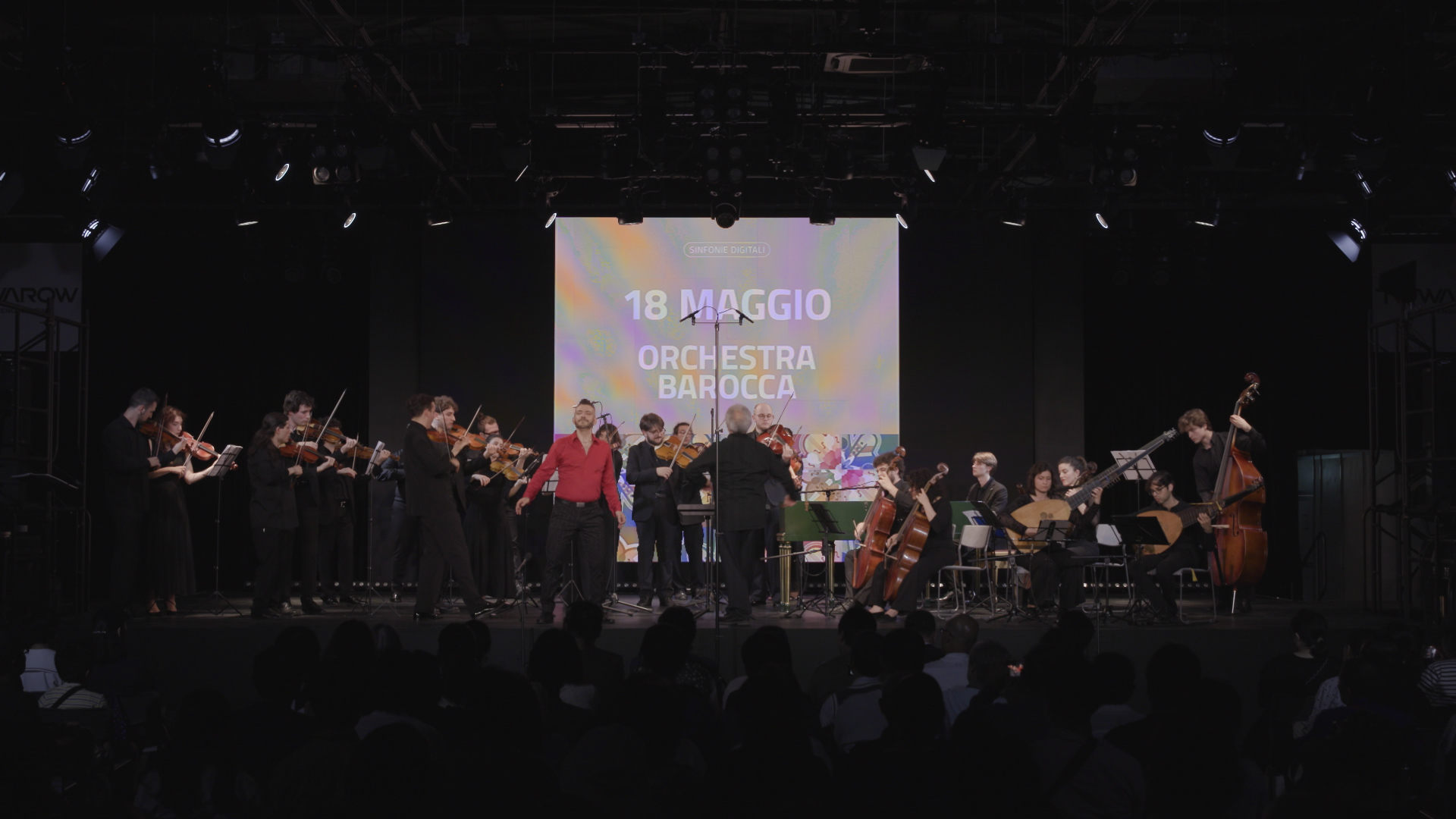 Image of live performance of an orchestra concert featuring the "orchestra barocca" on may 18. the musicians, dressed in formal attire, play various string instruments on a dimly lit stage with a large projection displaying the event title in the background.