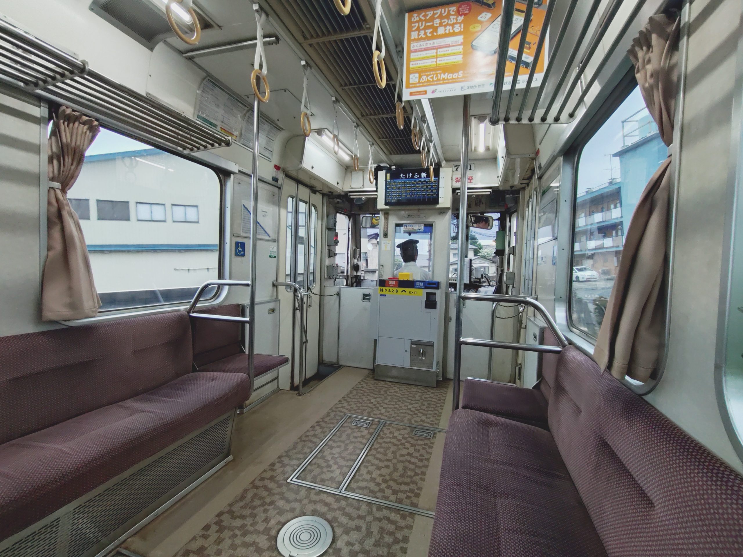 Image of interior of a japanese train in fukui, showcasing empty maroon seats, metal handrails, and safety features. the train's bright, well-organized interior is highlighted, offering a clear view of the urban landscape through large windows. public transportation,