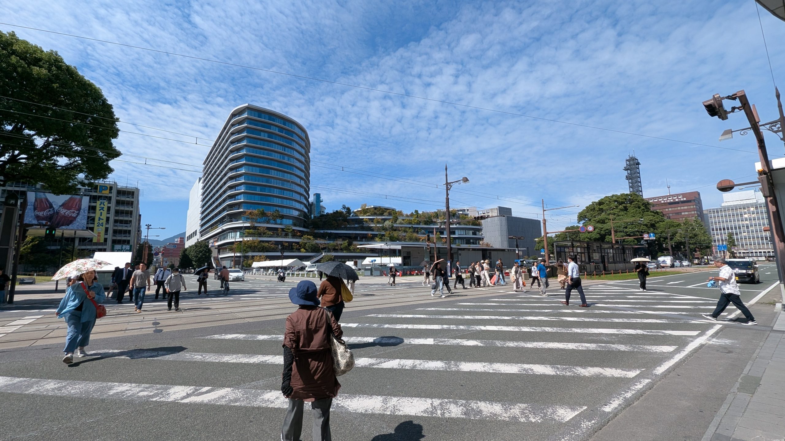 Image of a bustling urban scene in fukuoka, japan, featuring pedestrians crossing a wide street with modern architecture in the background. the distinctive multi-level fukuoka city library building, known for its innovative design, rises prominently under a clear, blue sky.